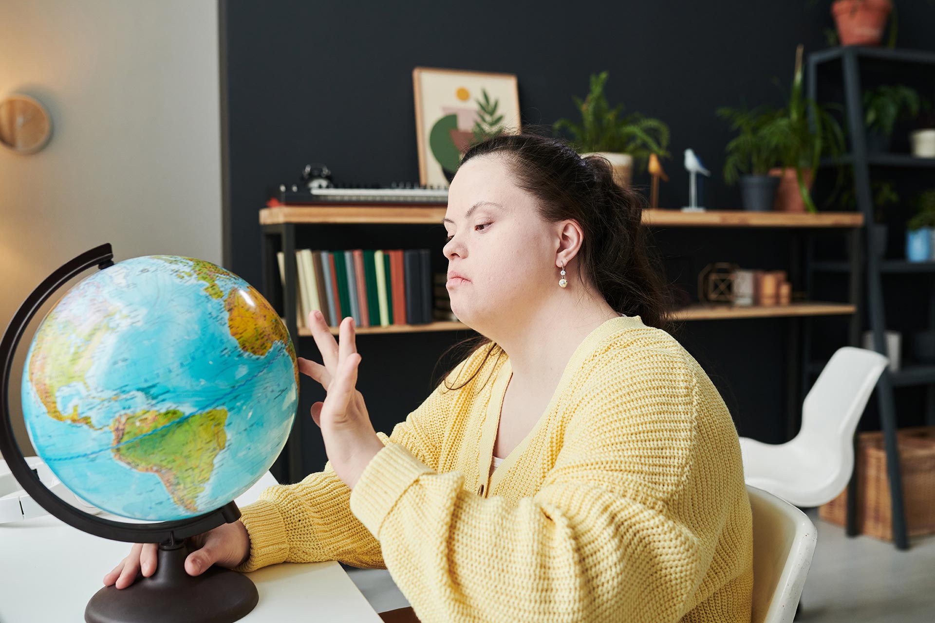 A woman with Down syndrome examining a globe in a cozy room filled with books and plants.